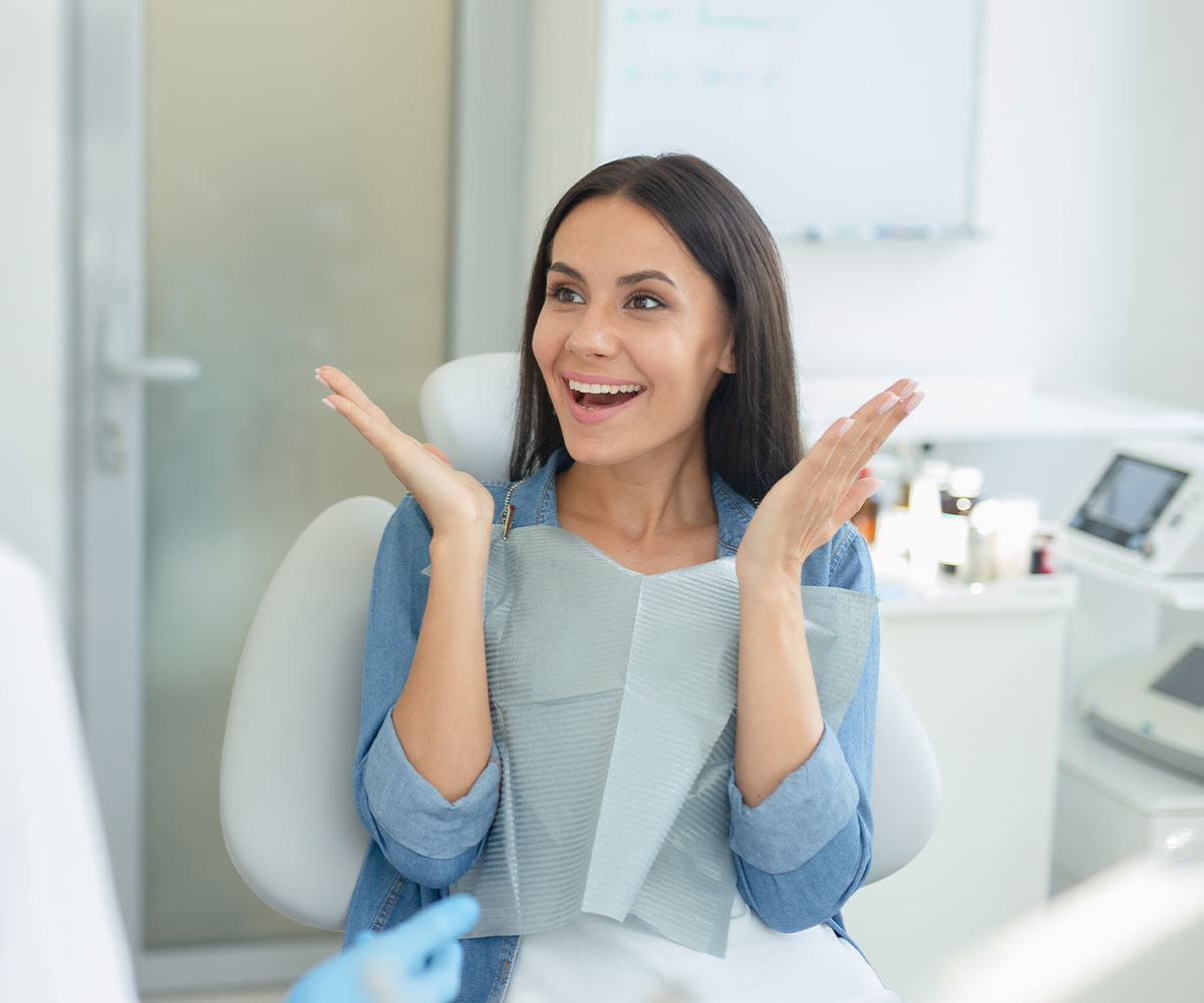 A smiling woman in a dental office, wearing a face mask and holding her hands up to her cheeks in surprise or excitement.